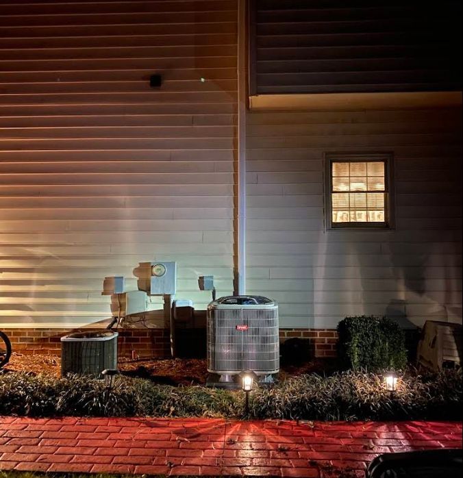 Exterior of a house at night with utility meters, bushes, and a lit window above a brick path