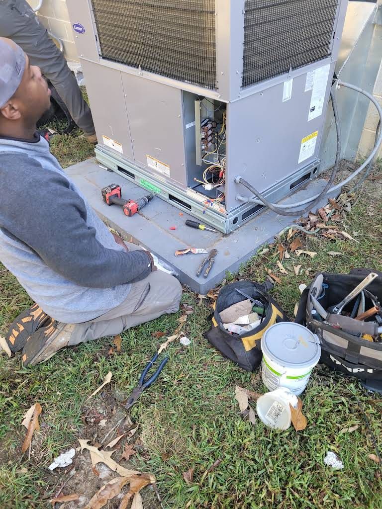 Technician kneeling by a large outdoor HVAC unit, with tools and paint supplies on the grass.