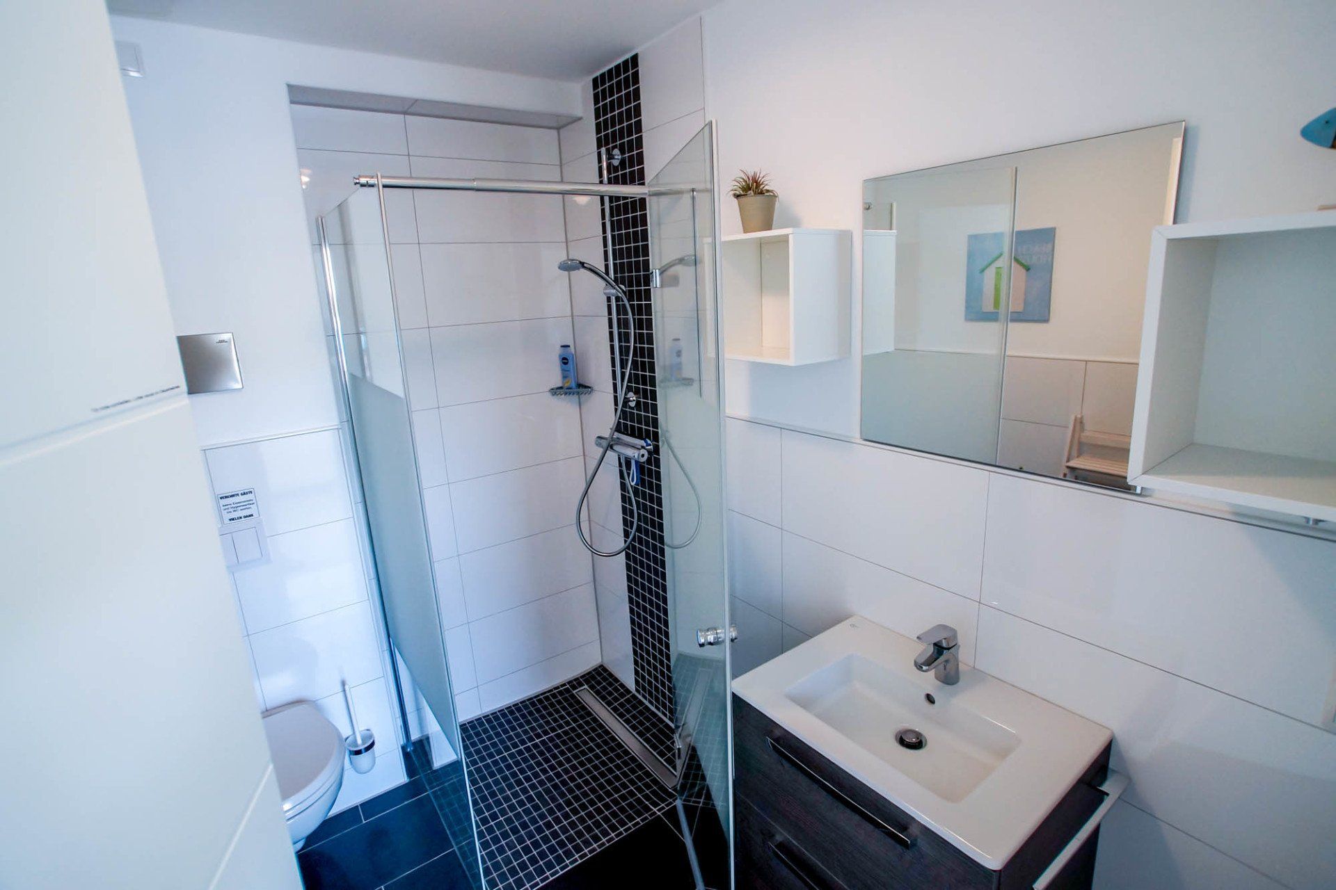 Kleines Badezimmer mit Dusche, Waschbecken, Spiegel und weiß gefliesten Wänden. Schwarz-weißer Mosaikboden.