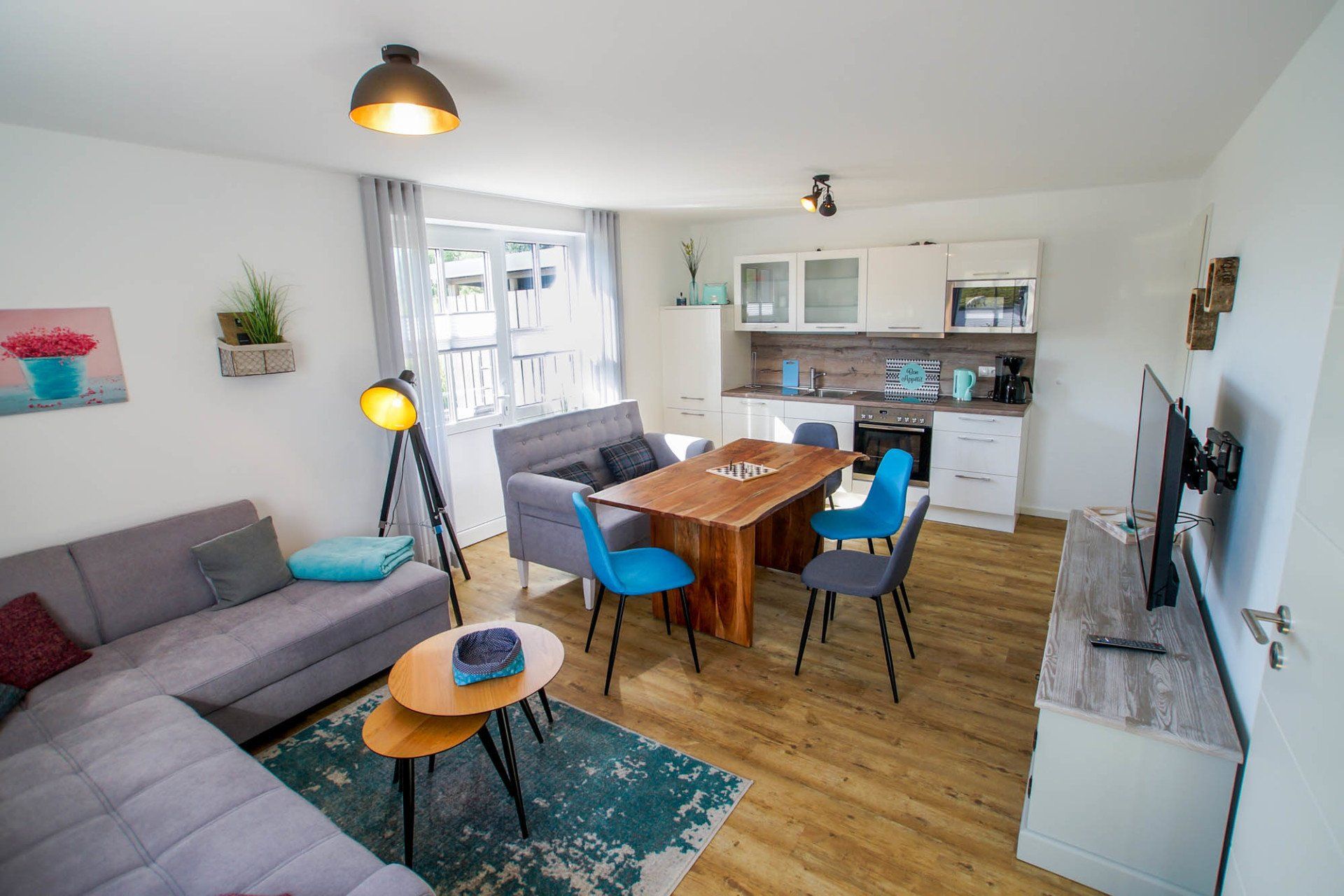 Moderner, offener Wohnbereich mit grauem Sofa, Esstisch, Küche und Fernseher. Hell, luftig und einladend.