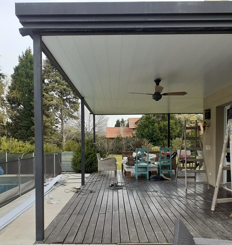 Patio cubierto de madera con mesa de comedor, sillas color turquesa y ventilador de techo, junto a una piscina y un jardín.