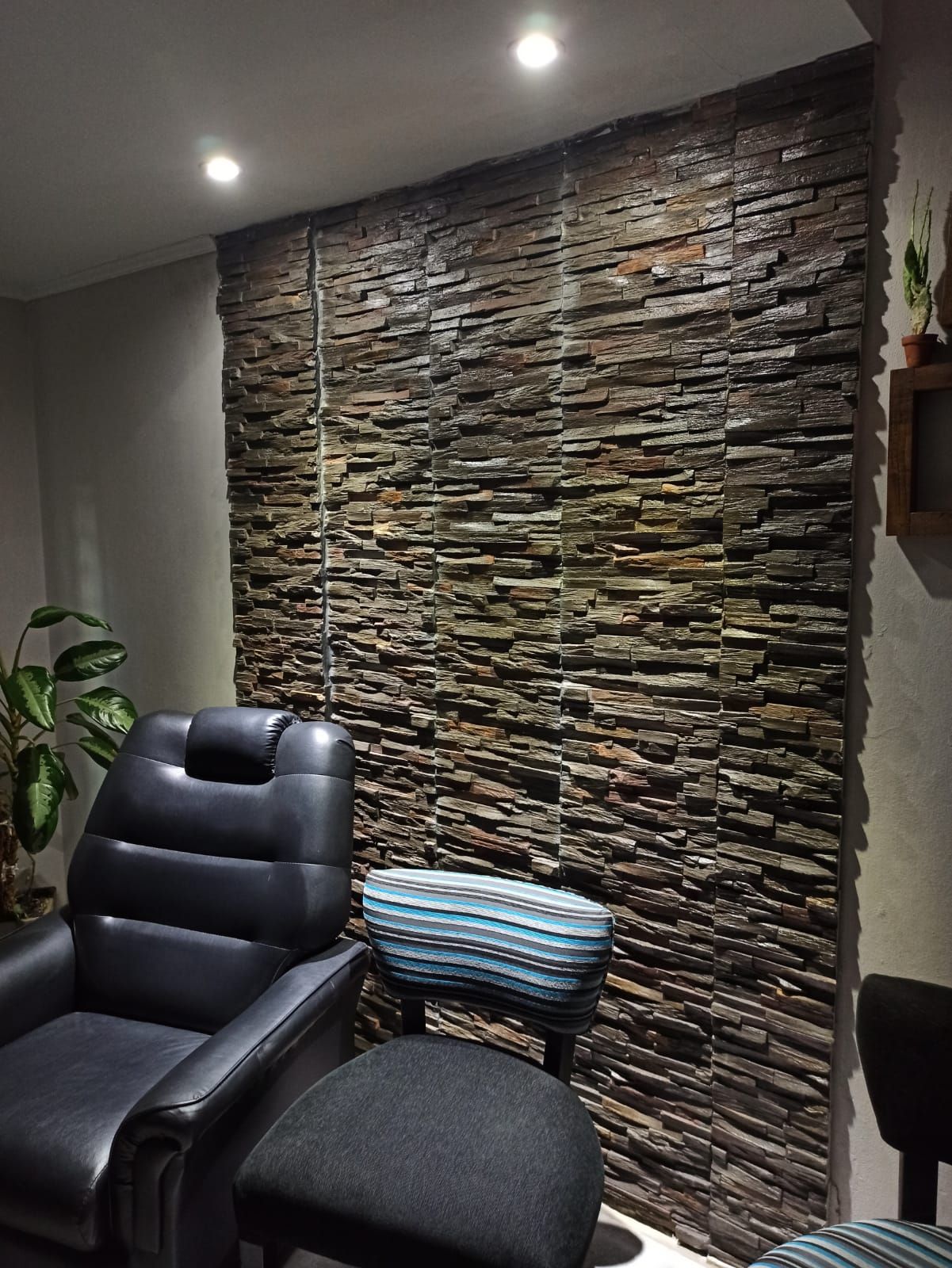 Pared decorativa de piedra detrás de un sillón reclinable negro en una habitación con poca luz, con una planta