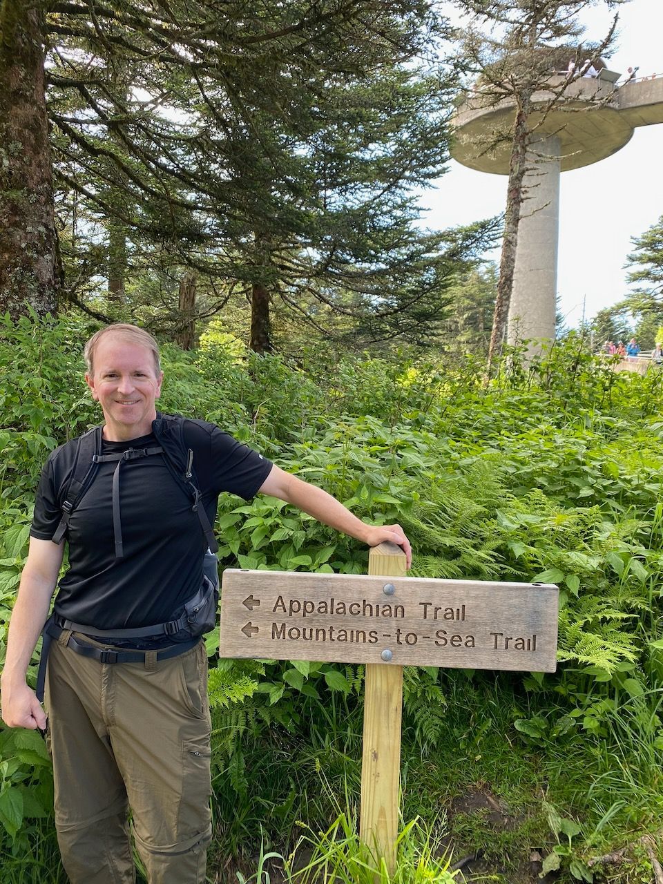 Person standing beside a sign for the “Abandoned Tree” and “Worms-to-Burn Trail” in a wooded area.