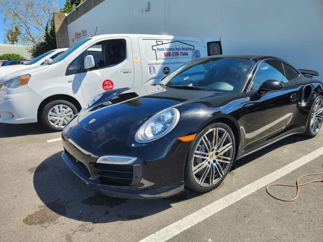 Black Porsche sports car parked next to a white van with business signage in a parking lot.