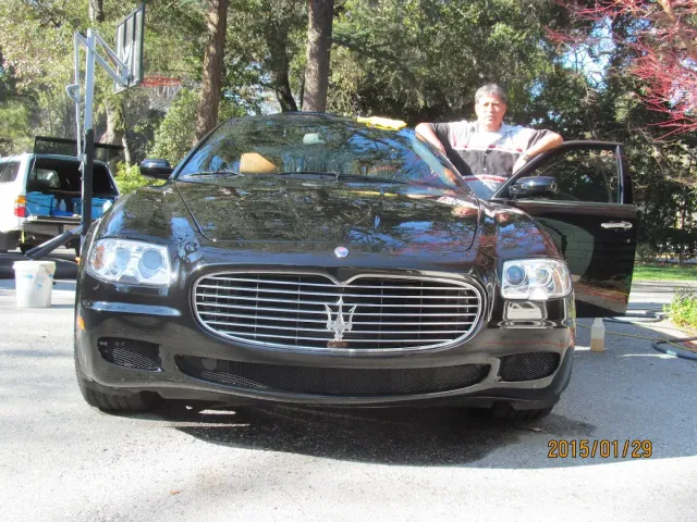 Man leaning on black Maserati in a driveway. Car is shiny, door open, trees in background.