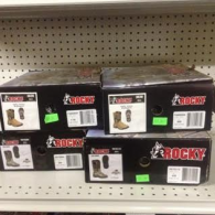 Four boxed pairs of Rocky brand work boots stacked on a store shelf, each showing product images and size labels.