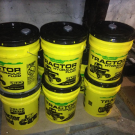 Six yellow 5-gallon buckets of 303 Tractor Hydraulic Fluid stacked in an indoor space.