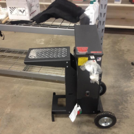 A portable, black metal log splitter with a diamond-plate platform, sitting on wheels in a warehouse setting.