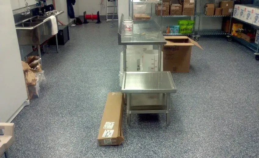 Commercial kitchen with stainless steel prep tables, boxes on the floor, and a gray speckled floor.