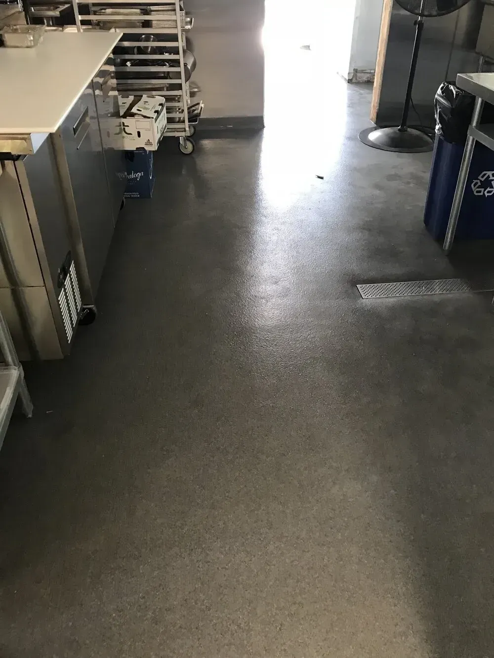 Commercial kitchen floor with stainless steel surfaces and equipment; bright sunlight through doorway.