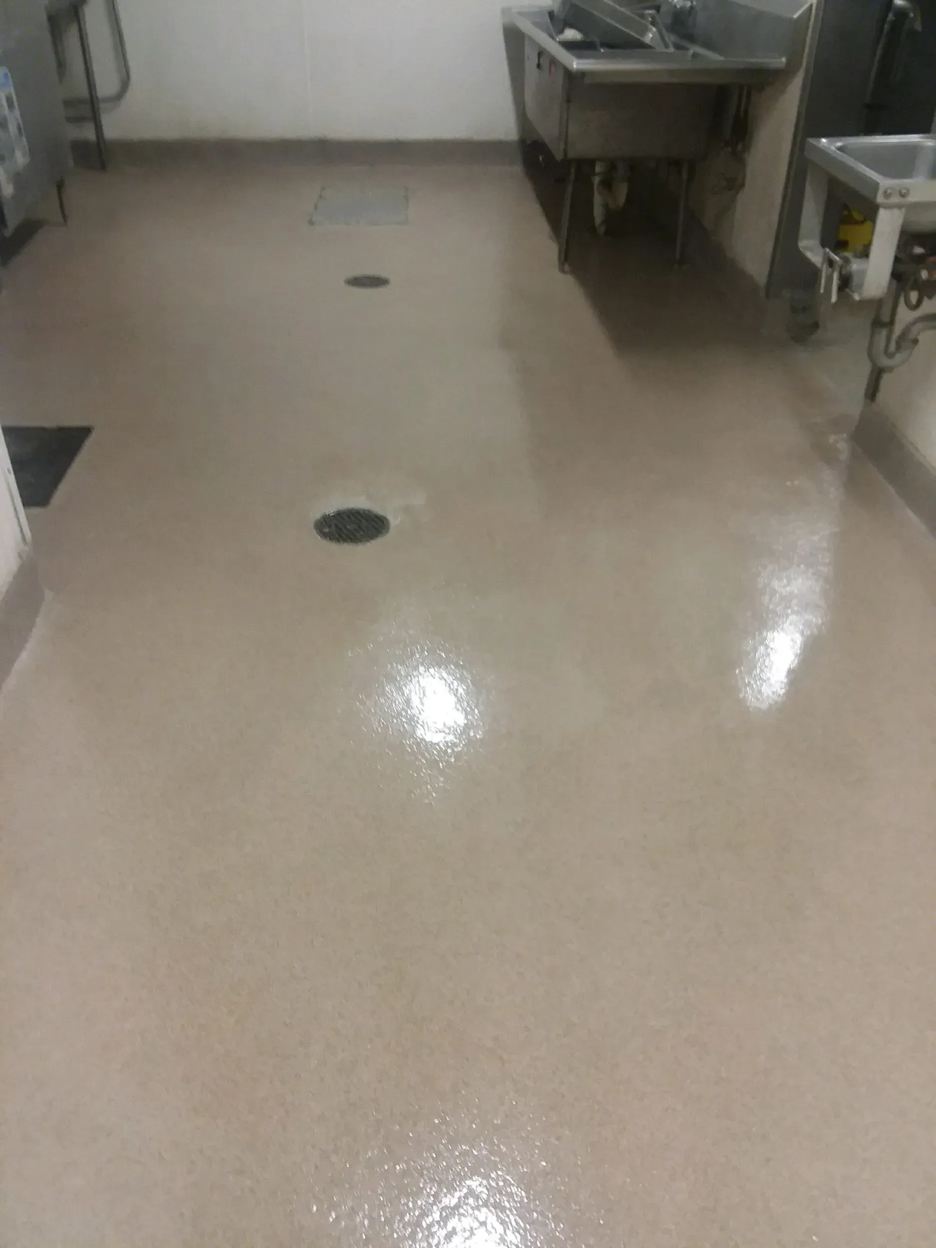 Tan, wet commercial kitchen floor with several drain covers, near a stainless steel sink.