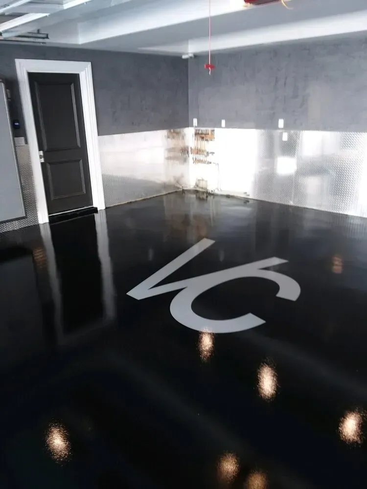 Black epoxy garage floor with silver logo, stainless steel backsplash, and dark gray walls.