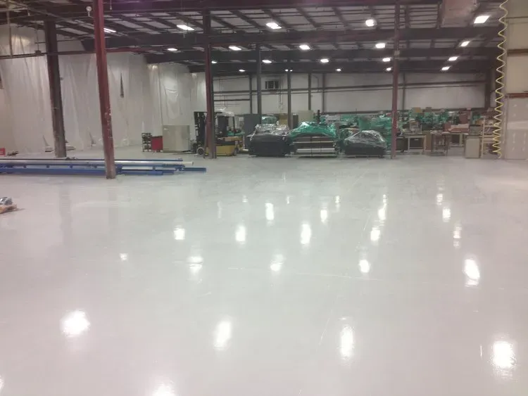 White, reflective industrial epoxy floor, with equipment and ceiling lights in a large warehouse.