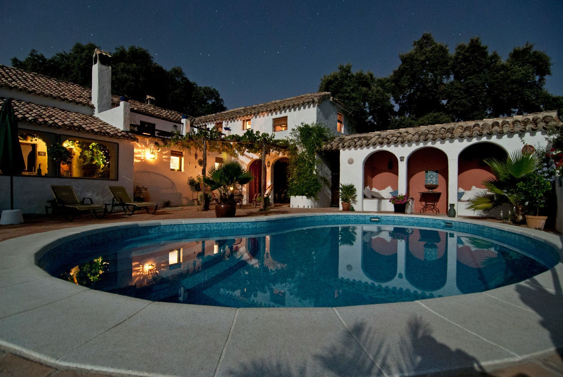 Nighttime shot of a villa with a round pool. Illuminated courtyard with arches, plants, and stars overhead.