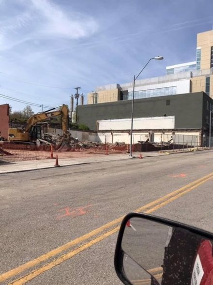 Construction site beside road with excavator, red dirt, and a large gray building under a blue sky