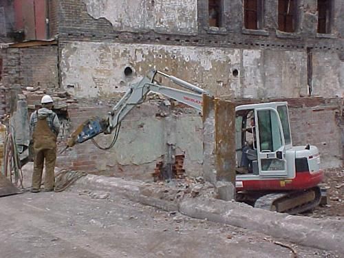 Excavator demolishing a damaged wall beside a worker in a rubble-strewn construction site