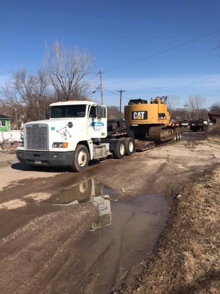 White tow truck hauling a yellow CAT machine on a muddy roadside under a clear blue sky