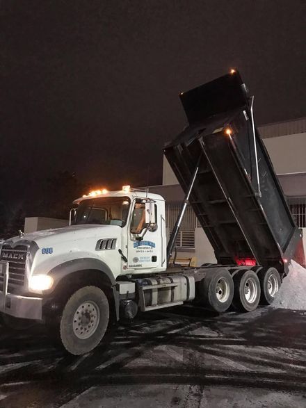 White dump truck with raised black bed on a snowy road at night, headlights and amber lights on