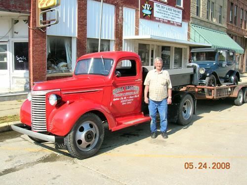 Red vintage pickup truck with trailer parked outside a storefront, with a man standing beside it.