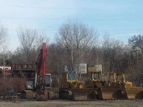 Heavy construction equipment, including excavators and bulldozers, parked in a dirt lot under bare trees.