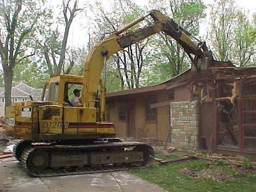 Yellow excavator demolishing a wooden building amid debris and trees
