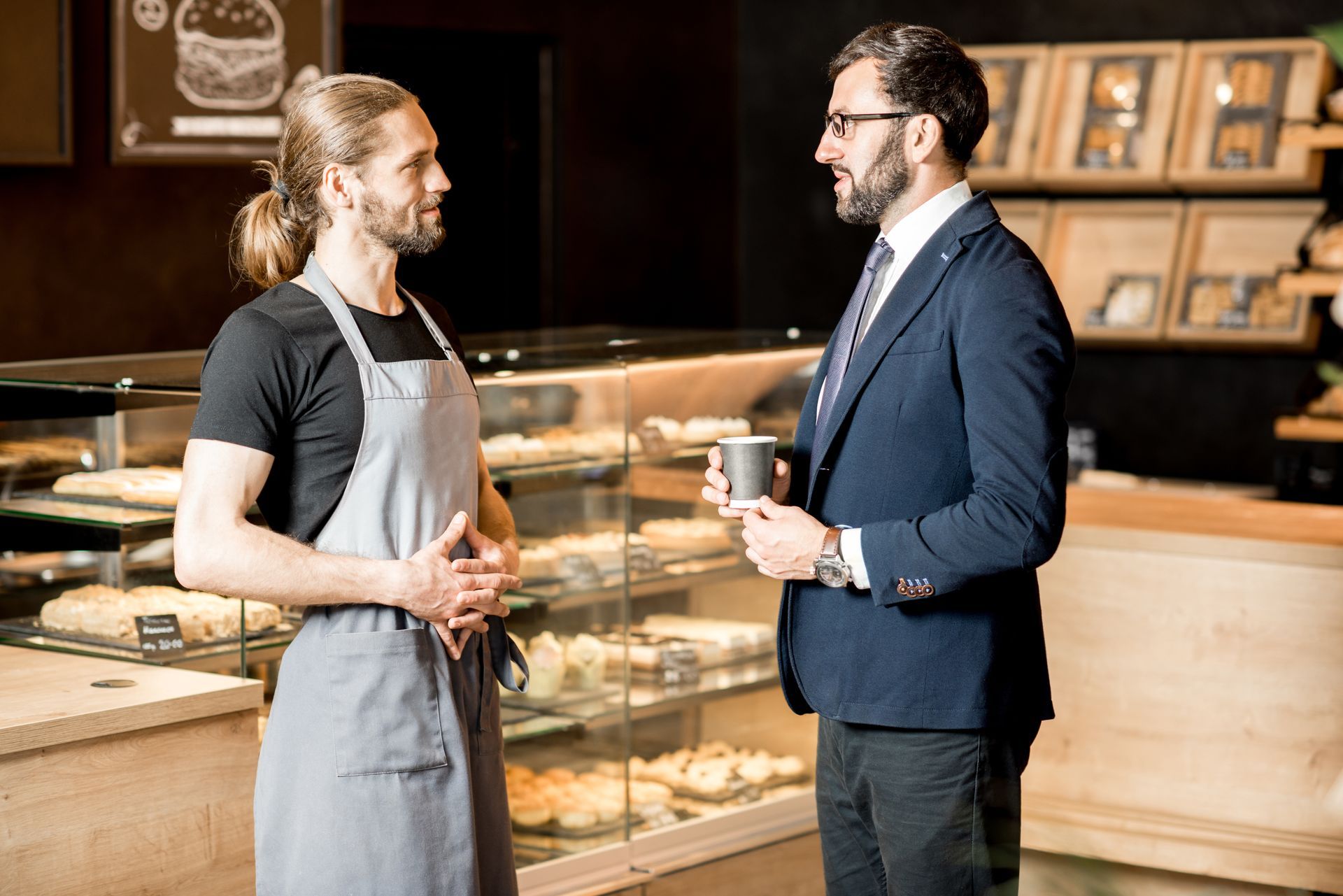 A man in a suit is talking to a man in an apron in a bakery.