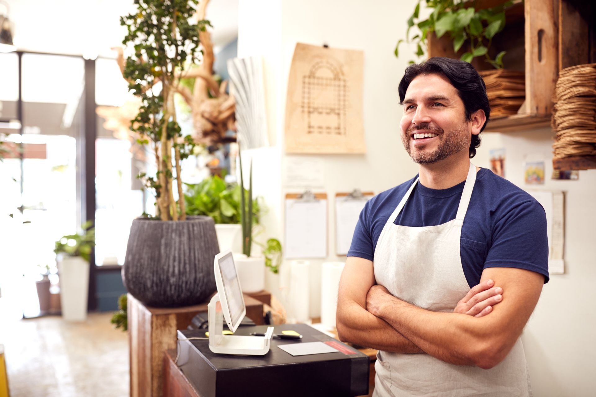 A man in an apron is standing in a restaurant with his arms crossed.
