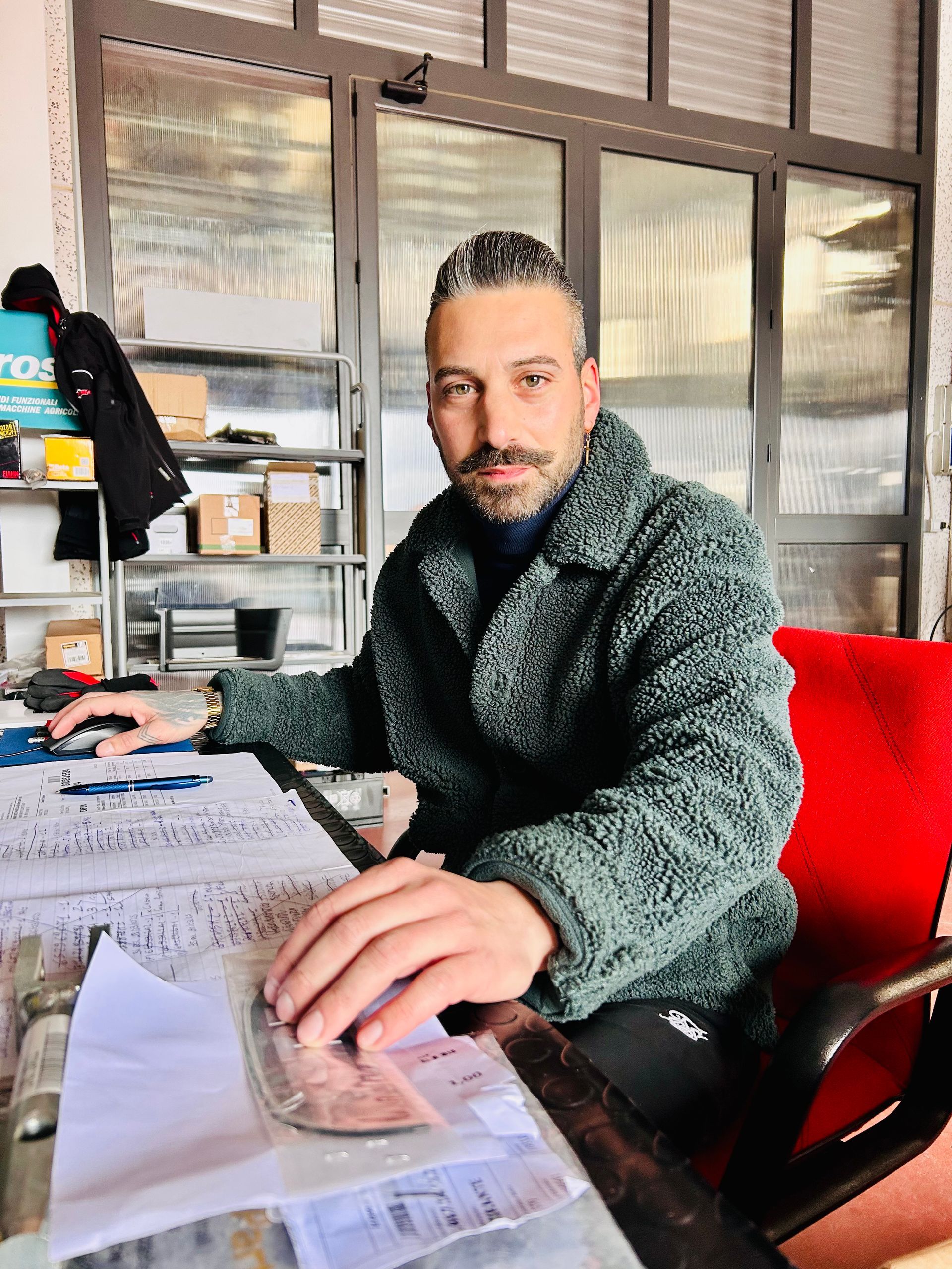 Un uomo in cappotto verde è seduto alla scrivania e guarda delle carte. Dietro di lui ci sono una finestra e degli scaffali.