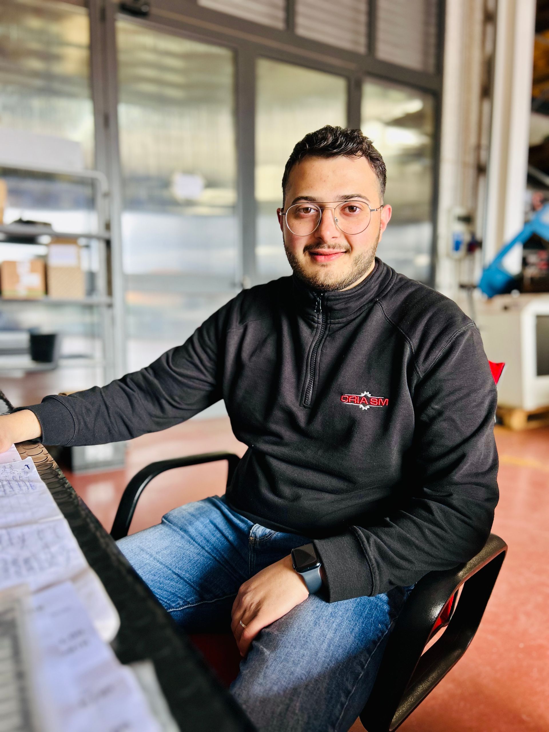 Uomo con occhiali, maglione nero e jeans seduto a una scrivania, sorridente. Ambiente industriale.