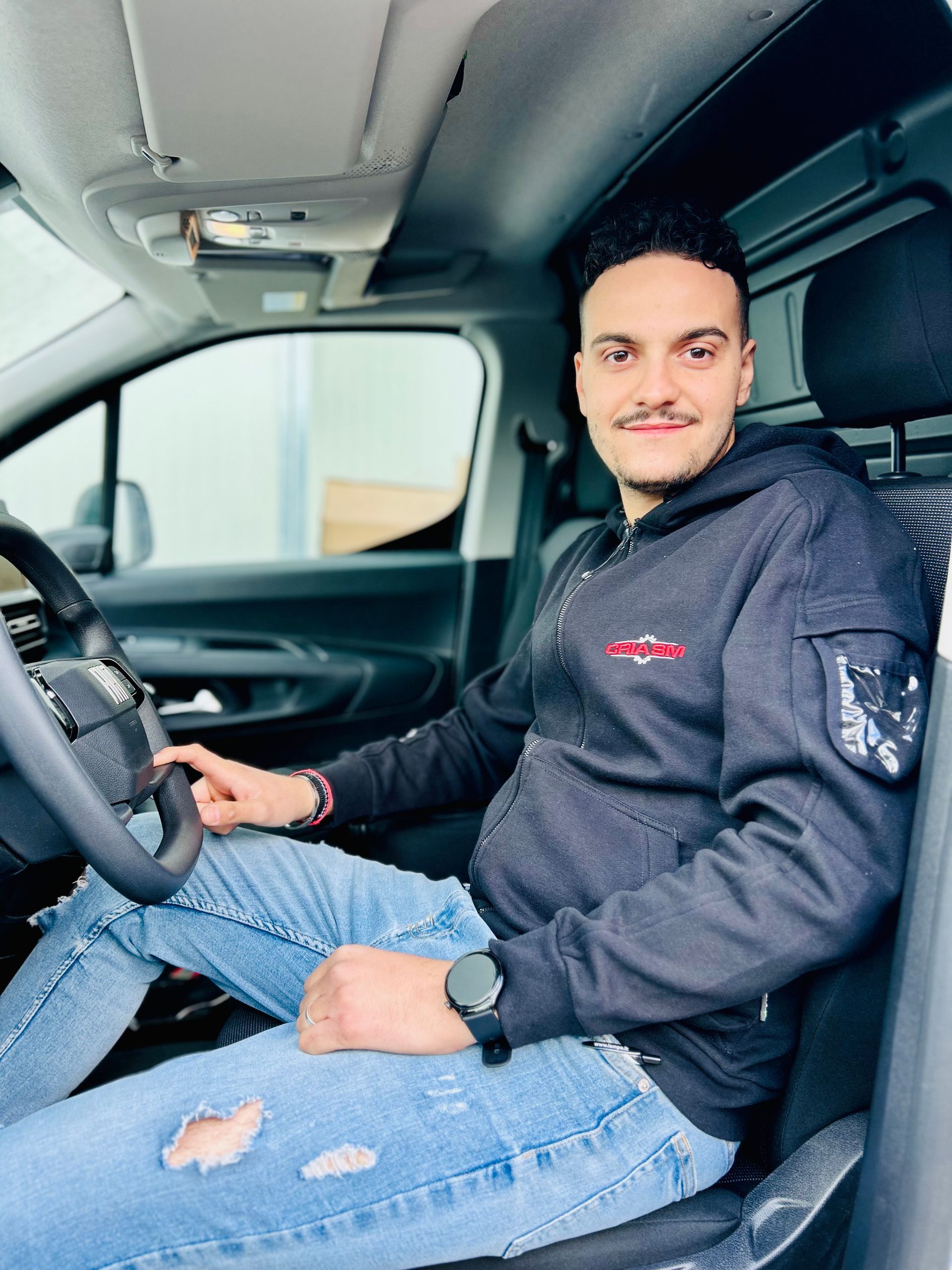 Un uomo con una giacca grigio scuro con cerniera e jeans strappati è seduto al posto di guida di un furgone bianco.