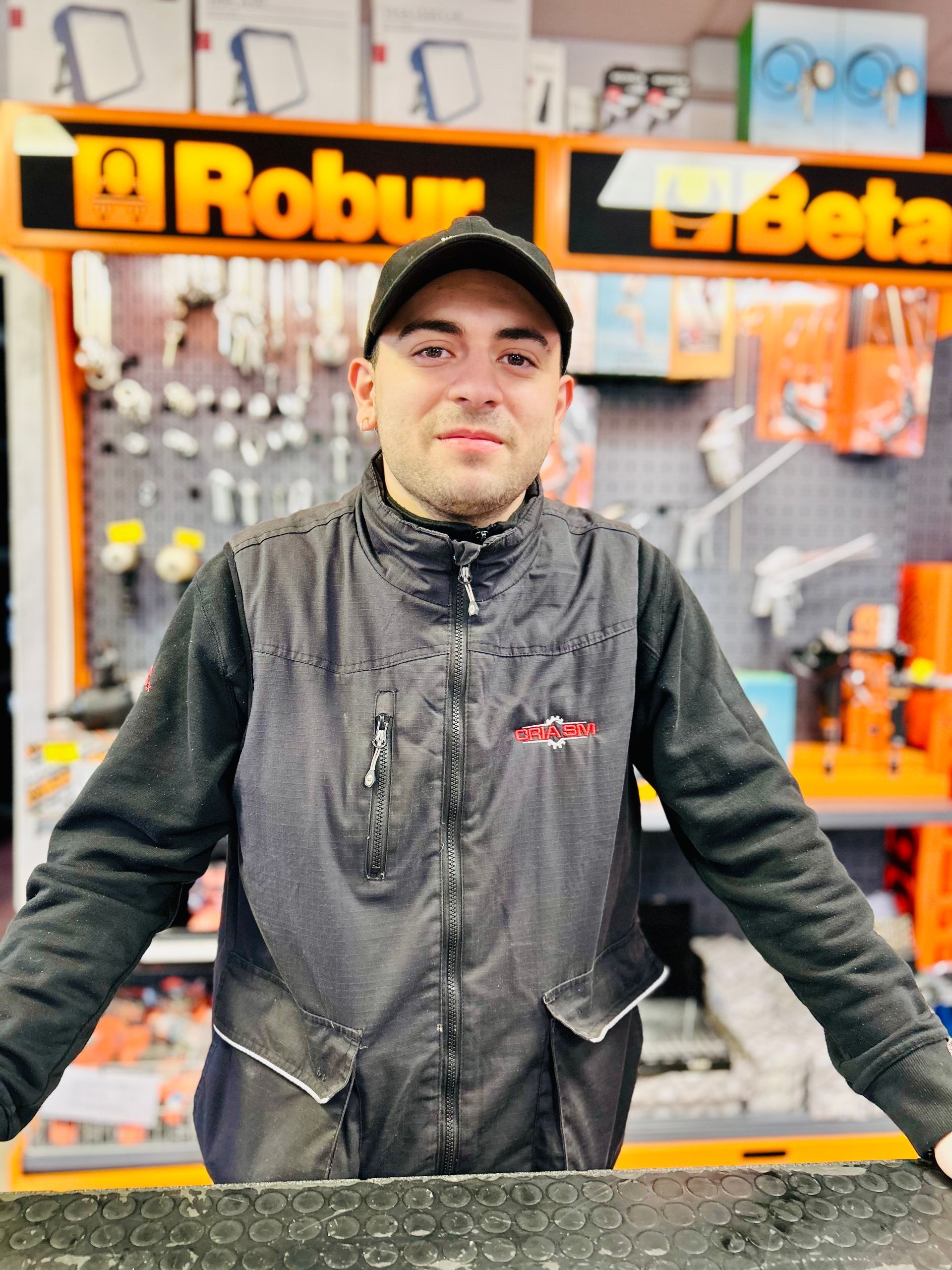 Un uomo con gilet e berretto neri, con le braccia tese, è in piedi in un negozio di ferramenta.