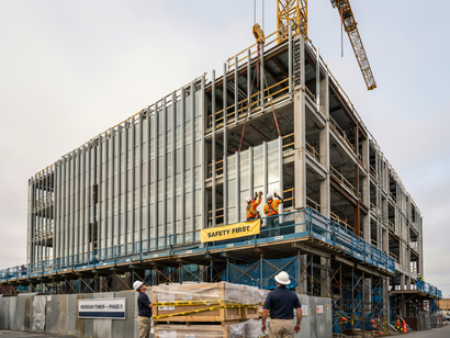 Construction site with a large steel-frame building under construction and workers on a blue platform