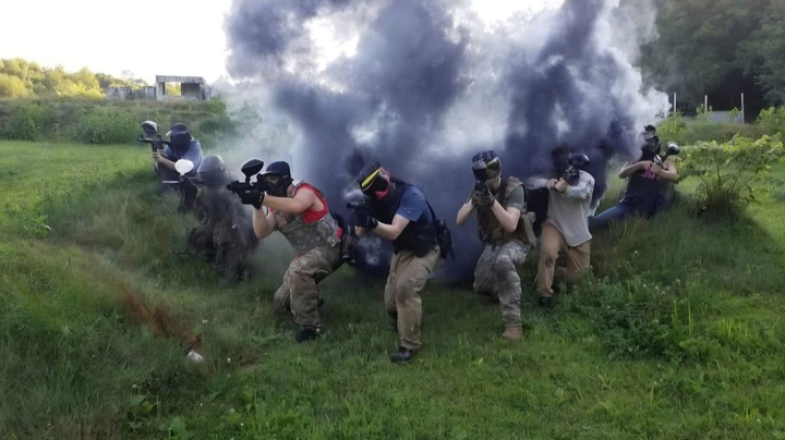 People in tactical gear crouch and move through grassy terrain as thick smoke billows behind them.