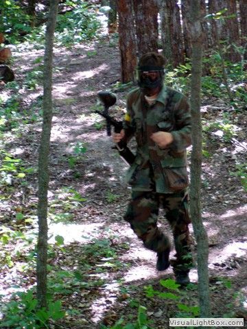 Camouflaged person holding a paintball marker walking through a wooded forest trail