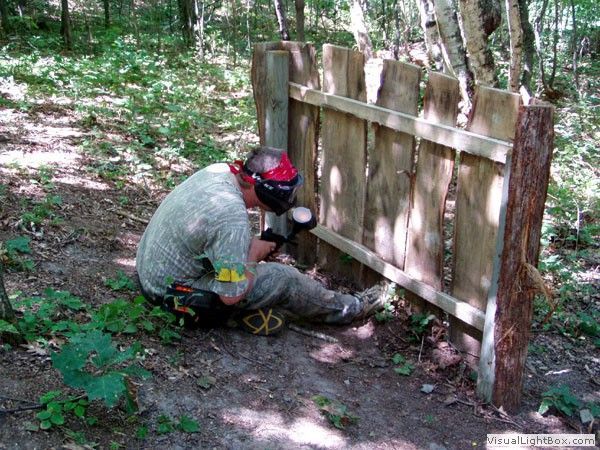 Person kneels by a wooden fence in a wooded area, using a power tool on the base of a post.