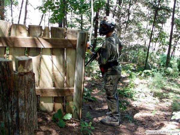 Person in camouflage stands by a wooden fence in a wooded outdoor area.