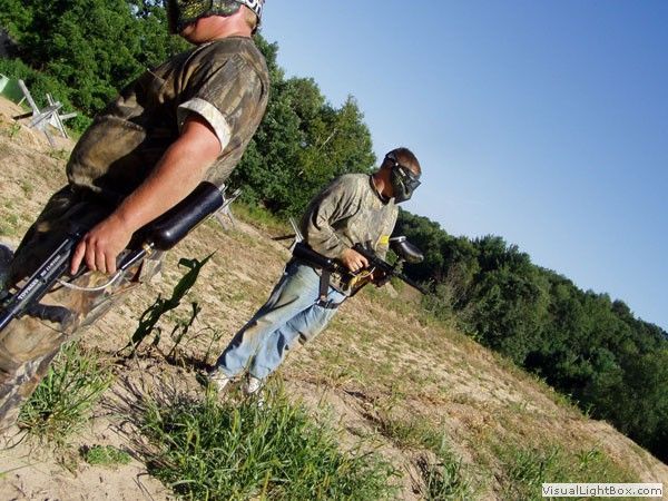 Two armed soldiers standing in a grassy field near trees, one in camouflage and one in a tan shirt.