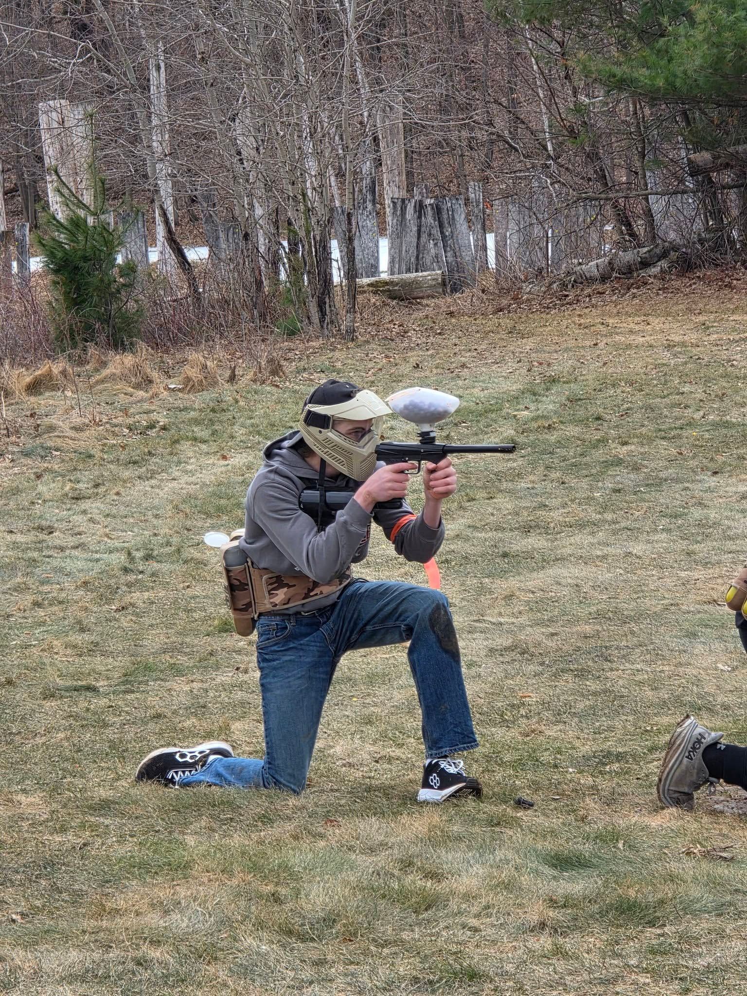 Person kneeling outdoors aiming a paintball marker, wearing a helmet and gray jacket in a grassy yard.
