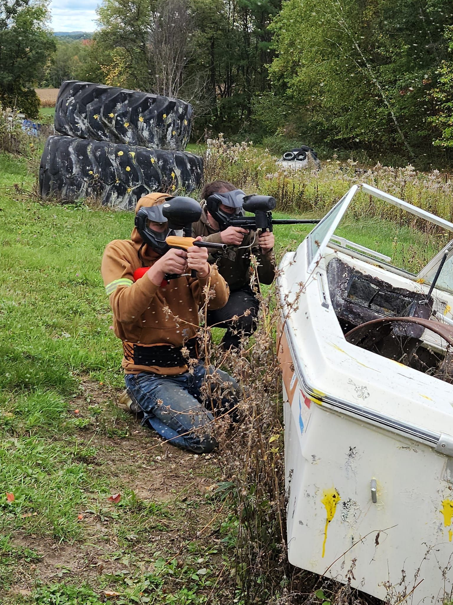 Two people kneel behind cover outdoors, aiming rifles toward a white vehicle in a grassy field.