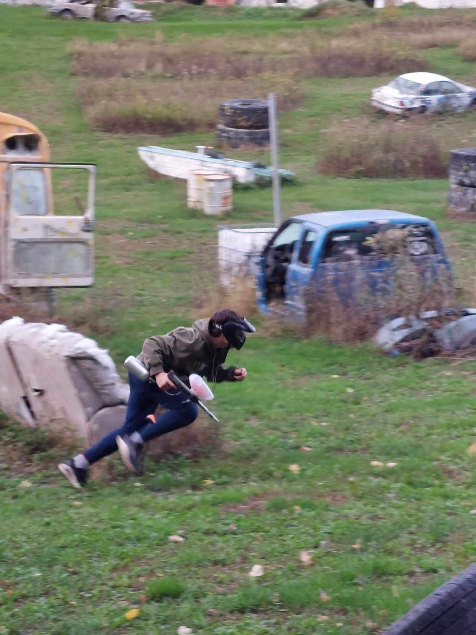 Person in dark clothing running with a paintball gun across a grassy outdoor field with barriers.