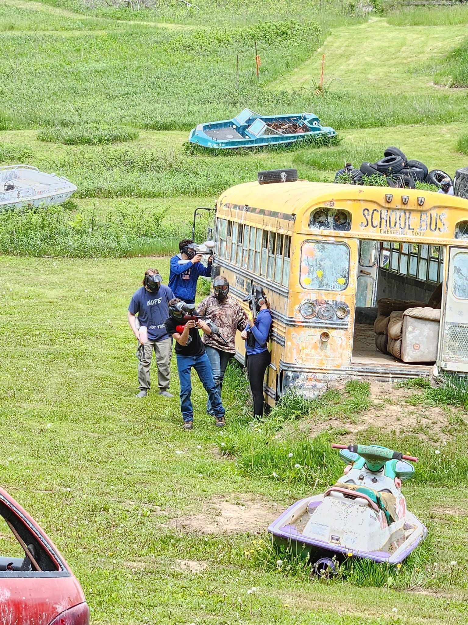 People near a yellow bus in a grassy field, with a blue car on the hill in the background.