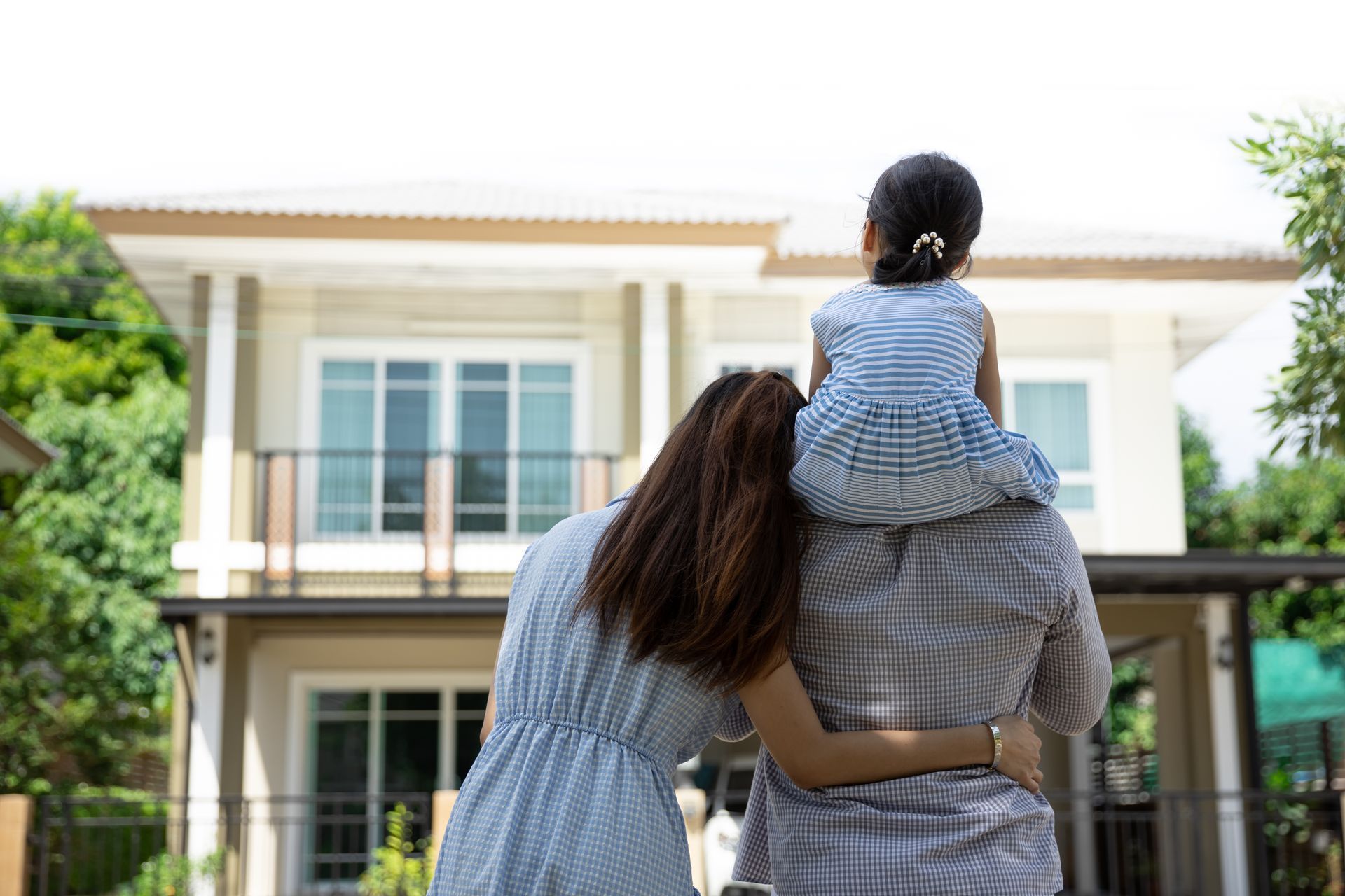 Adult carrying a child on shoulders in front of a modern house, viewed from behind.