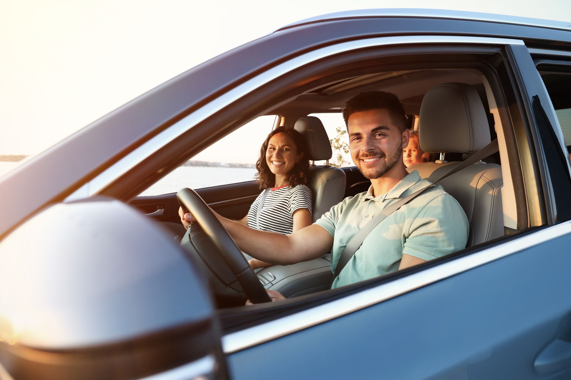 Three people sit in a car with seatbelts on, viewed through the open driver side window.