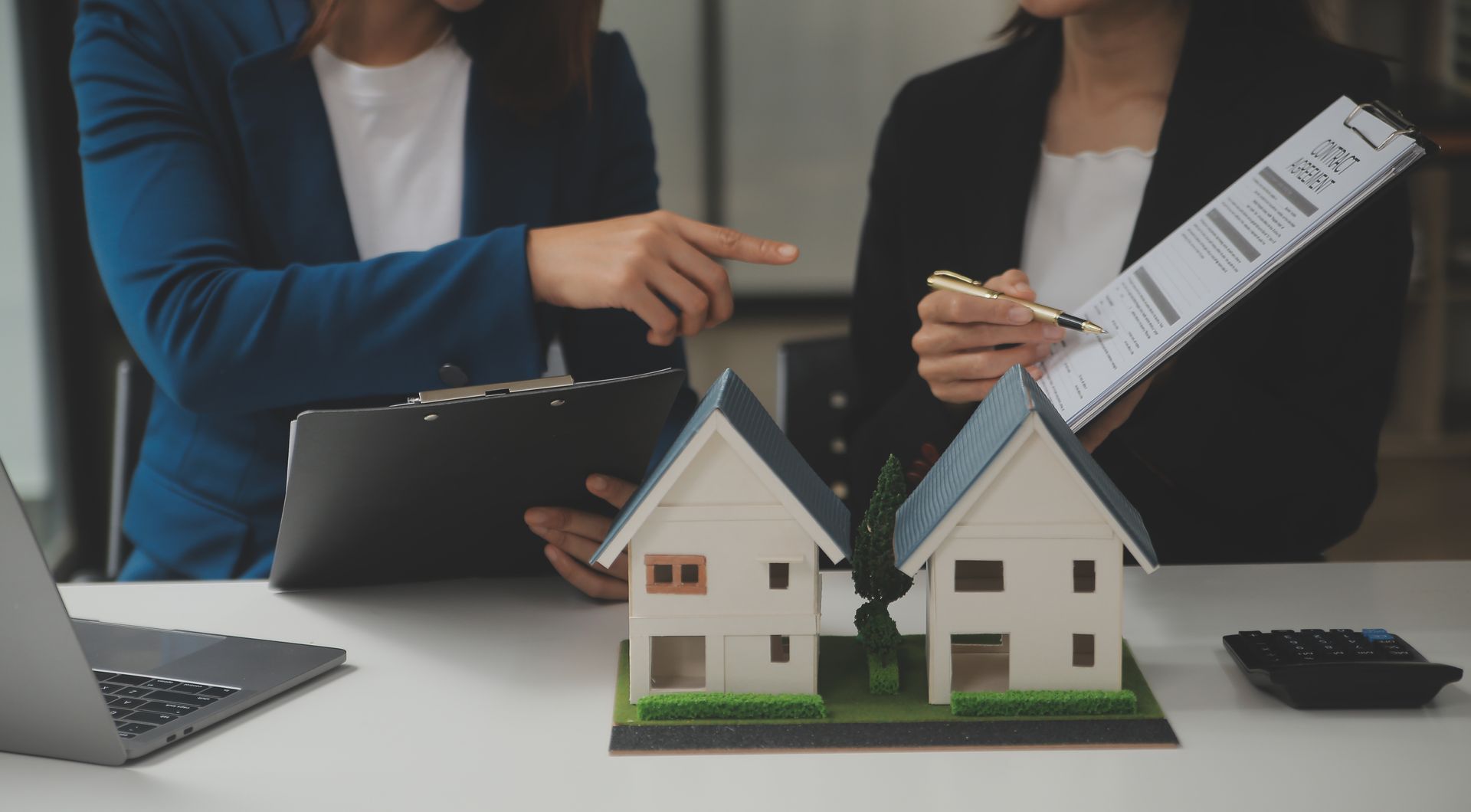 Two professionals reviewing a home insurance policy on a clipboard beside model houses on a desk