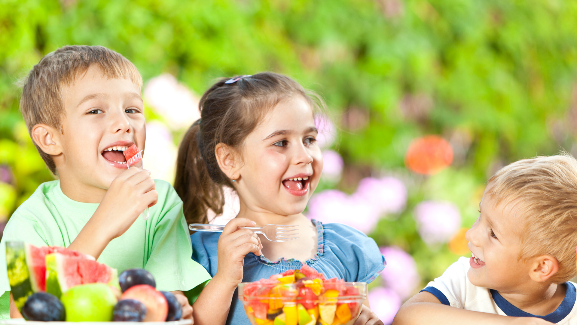 Three happy children eating fruit outdoors.