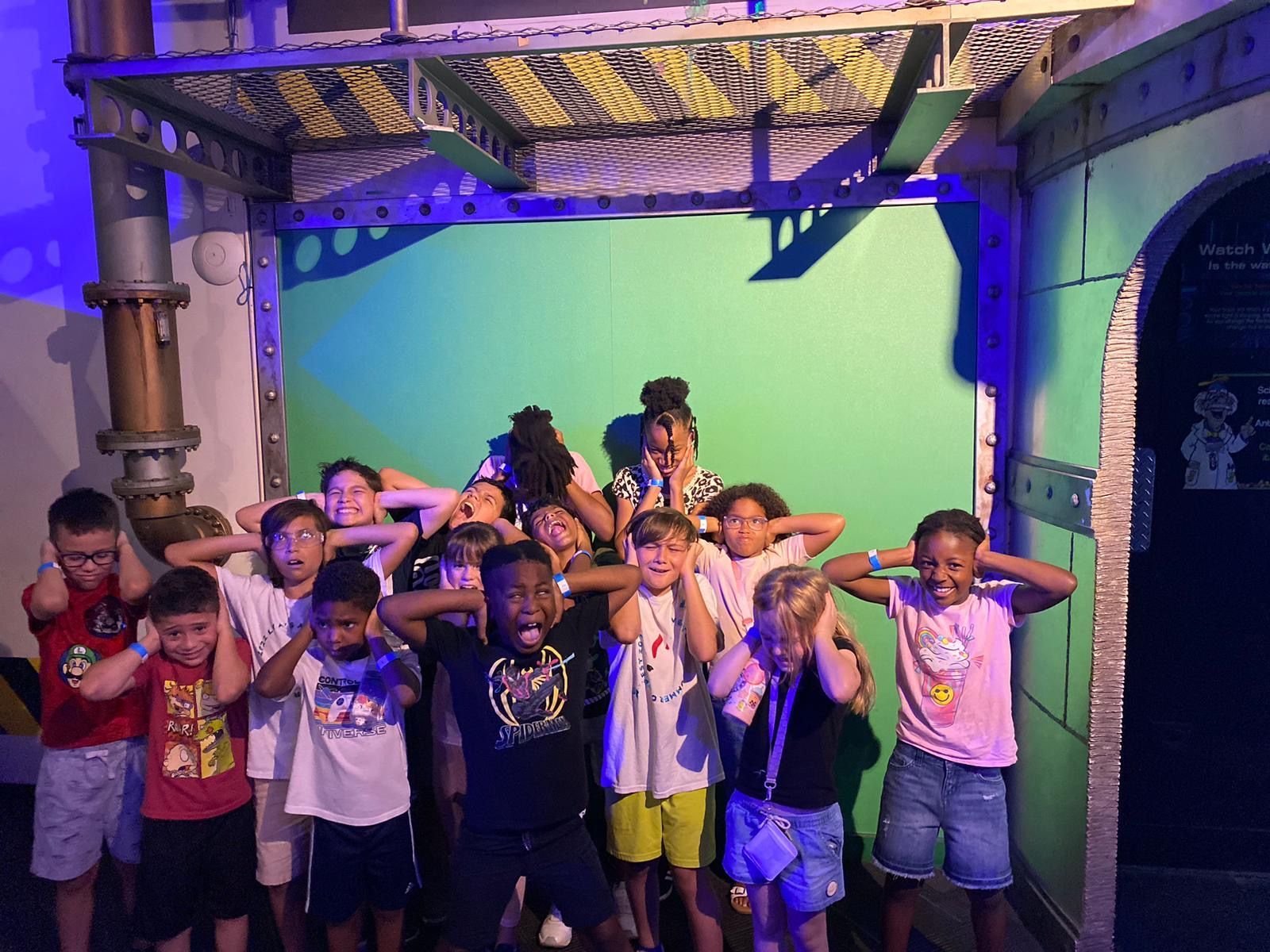 A group of children are standing in front of a green screen covering their ears.