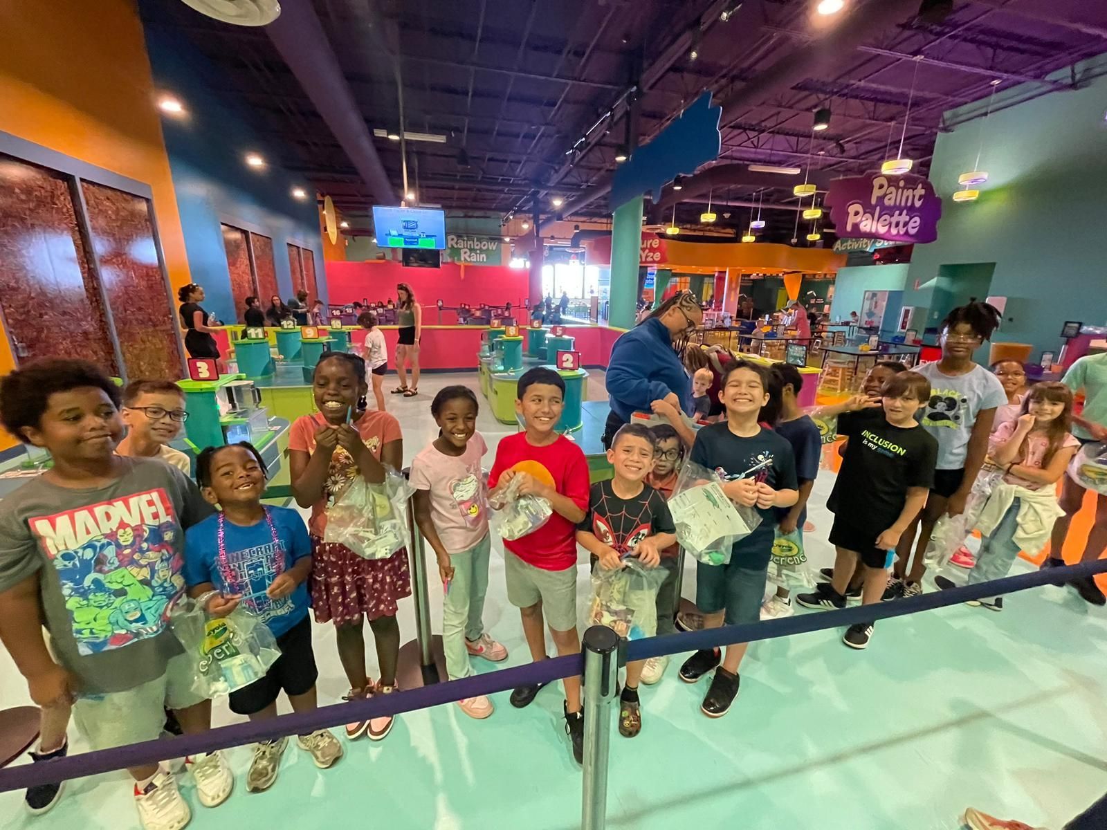 Group of diverse children smiling, holding bags at Crayola Experience; indoor space.