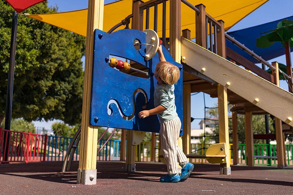 Two children sliding down a yellow slide on a playground; green turf, blue sky.