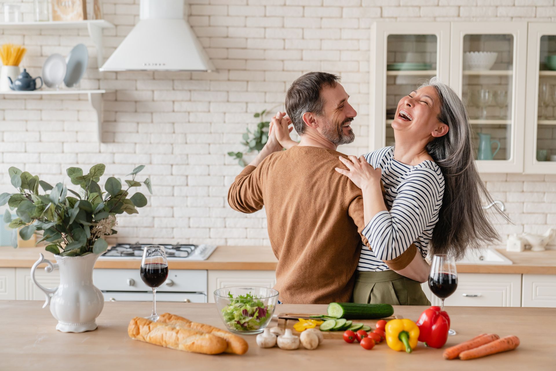 Couple laughing in a bright kitchen with fresh vegetables and bread on the counter