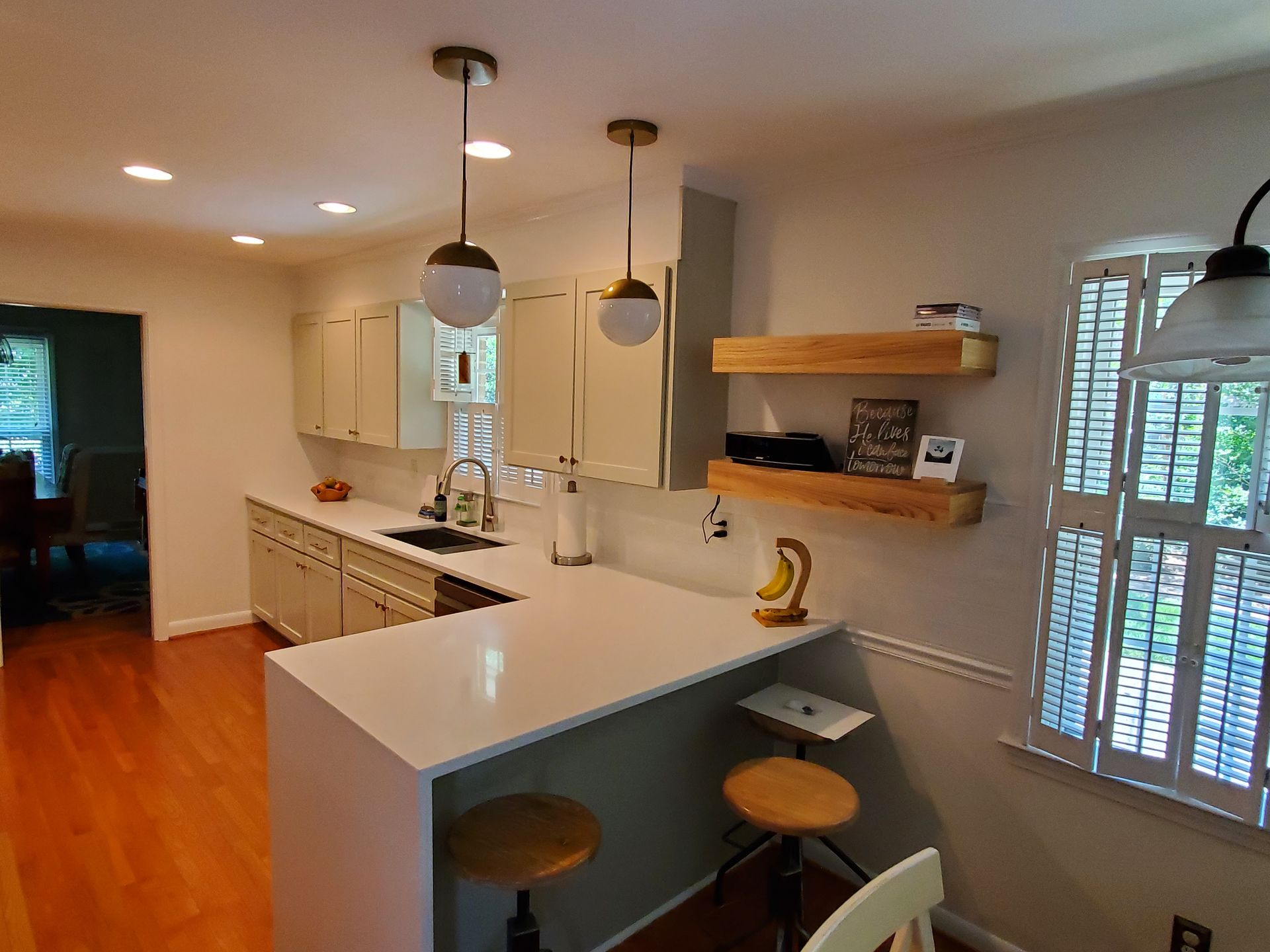 A kitchen with white cabinets , white counter tops , a sink , stools and a window.